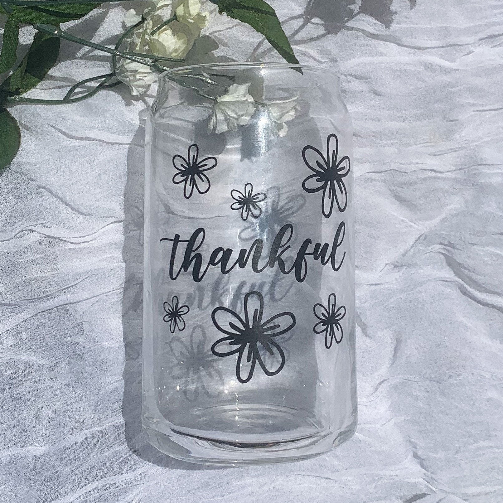 A glass can with thankful written on it in cursive and is surrounded by flowers.