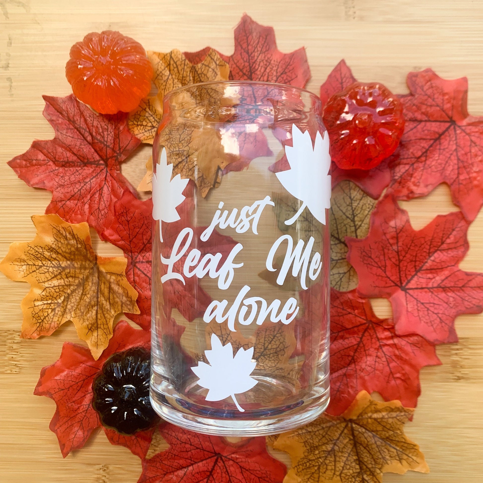Just leaf me alone fall glass cup surrounded by colorful leaves and mini pumpkins.