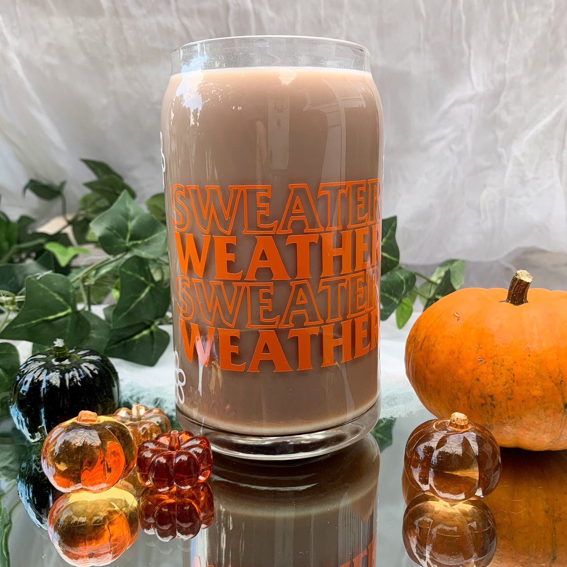 Sweater weather glass can sitting on a mirror with mini pumpkins in front of a white backdrop.